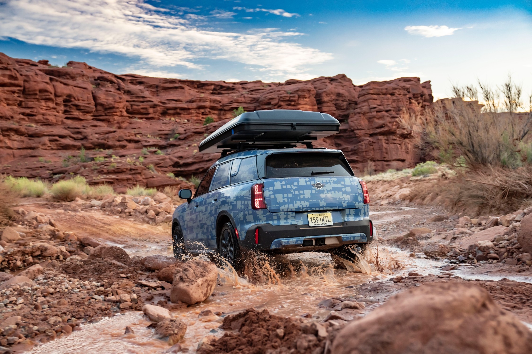 MINI Countryman visto pela traseira, a atravessar um riacho em terreno rochoso, com  uma caixa de tejadilho 